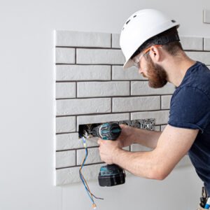 Electrician builder in a white helmet at work with a drill, installation of sockets, and switches.